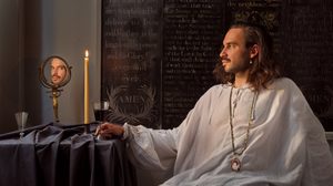 The Reflected Self. © Simon Bevan A man with long hair and a beard sits at a table in a dimly lit room. He's wearing a white loose-fitting shirt and a necklace with a pendant. On the table are a lit candle, a glass of dark liquid, and a small mirror. Behind him is a wall with religious text inscribed on it.