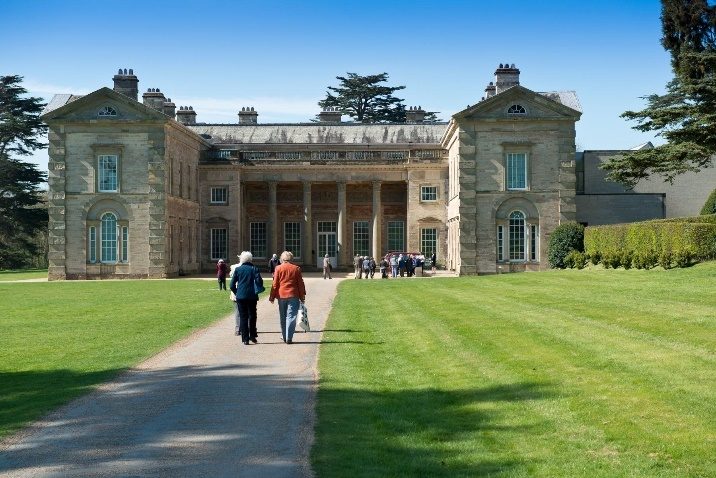 A Georgian manor house set in grassy grounds. People walk up a long gravel driveway towards the house.