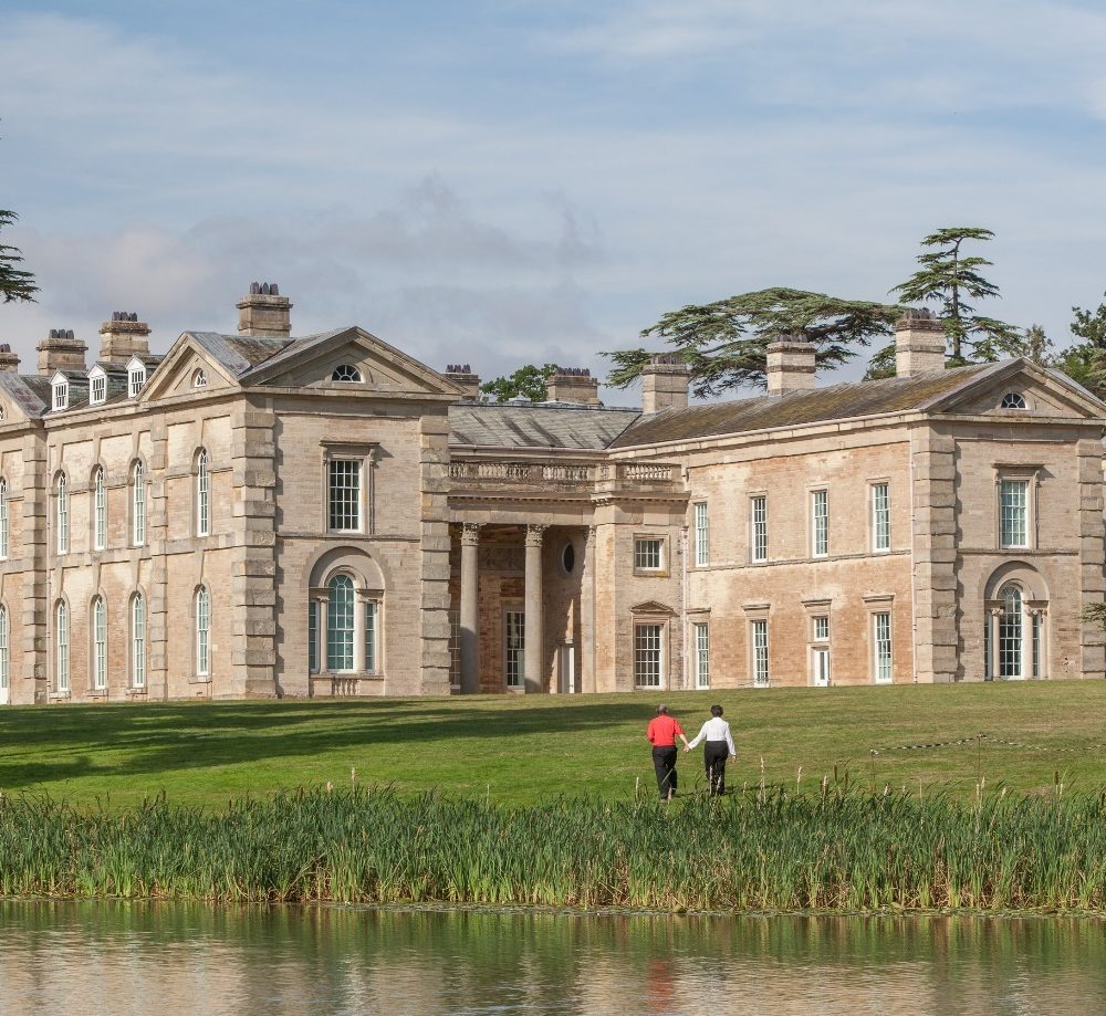 A Georgian manor house set in grassy grounds, surrounded by trees and a lake at the front. A couple holding hands walk across the grass towards the house.