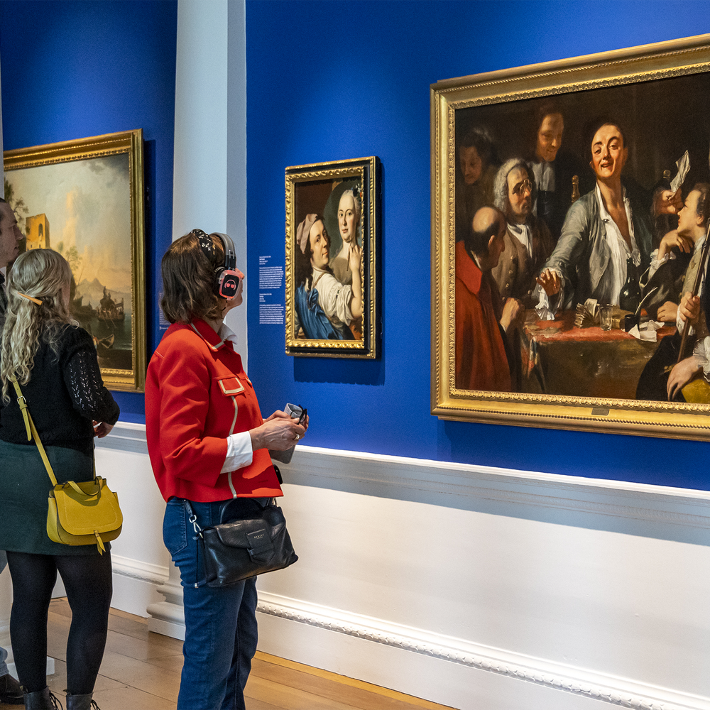 The image depicts a group of visitors at an art gallery, observing several framed paintings on a blue wall. The artworks appear to be portraits or genre scenes from the Baroque or Renaissance period, with ornate gold frames. The visitors, wearing casual modern attire, seem engrossed in studying the details of the paintings.