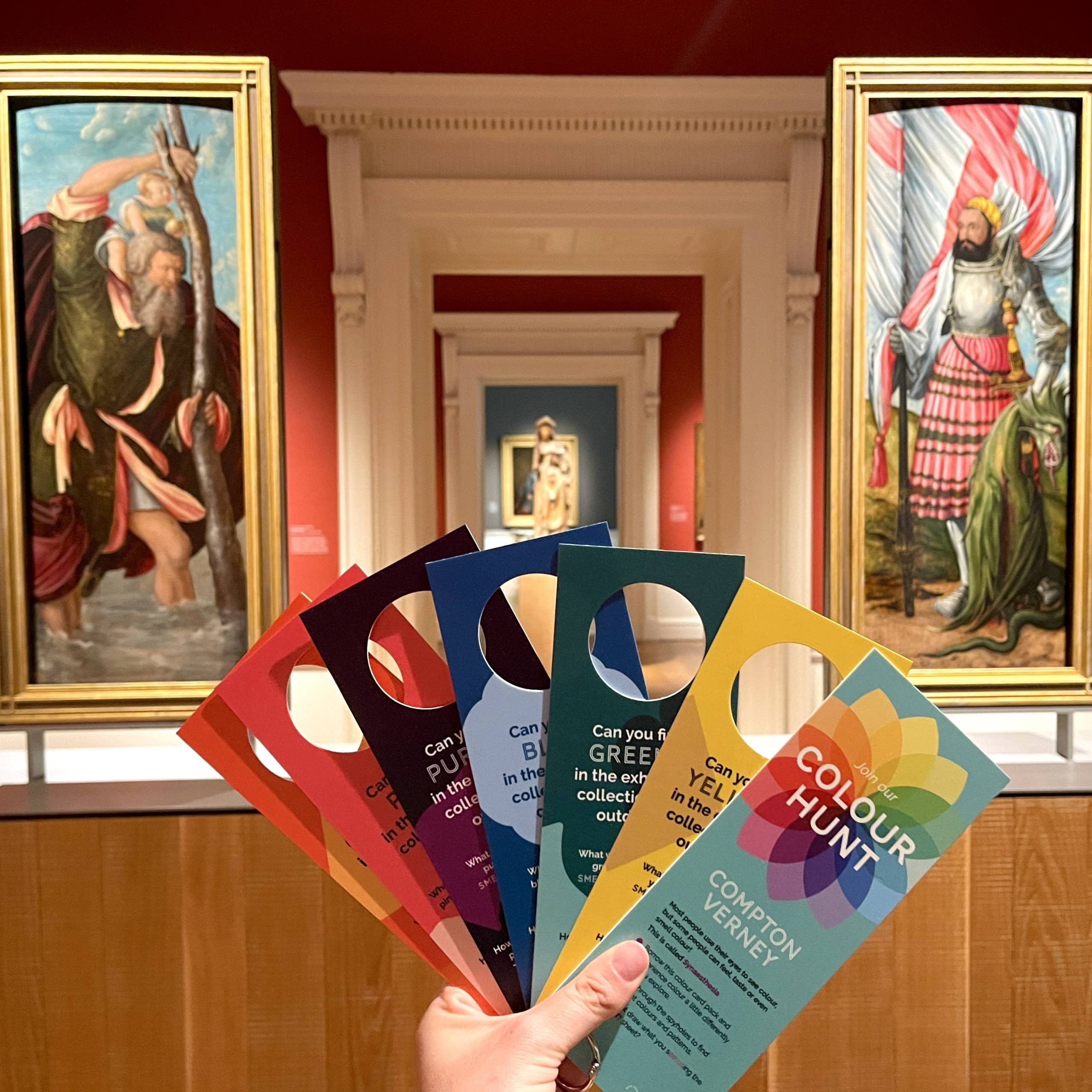 An interior gallery space at Compton Verney, featuring colourful framed paintings on the walls and a hand holding a fan of cards titled 