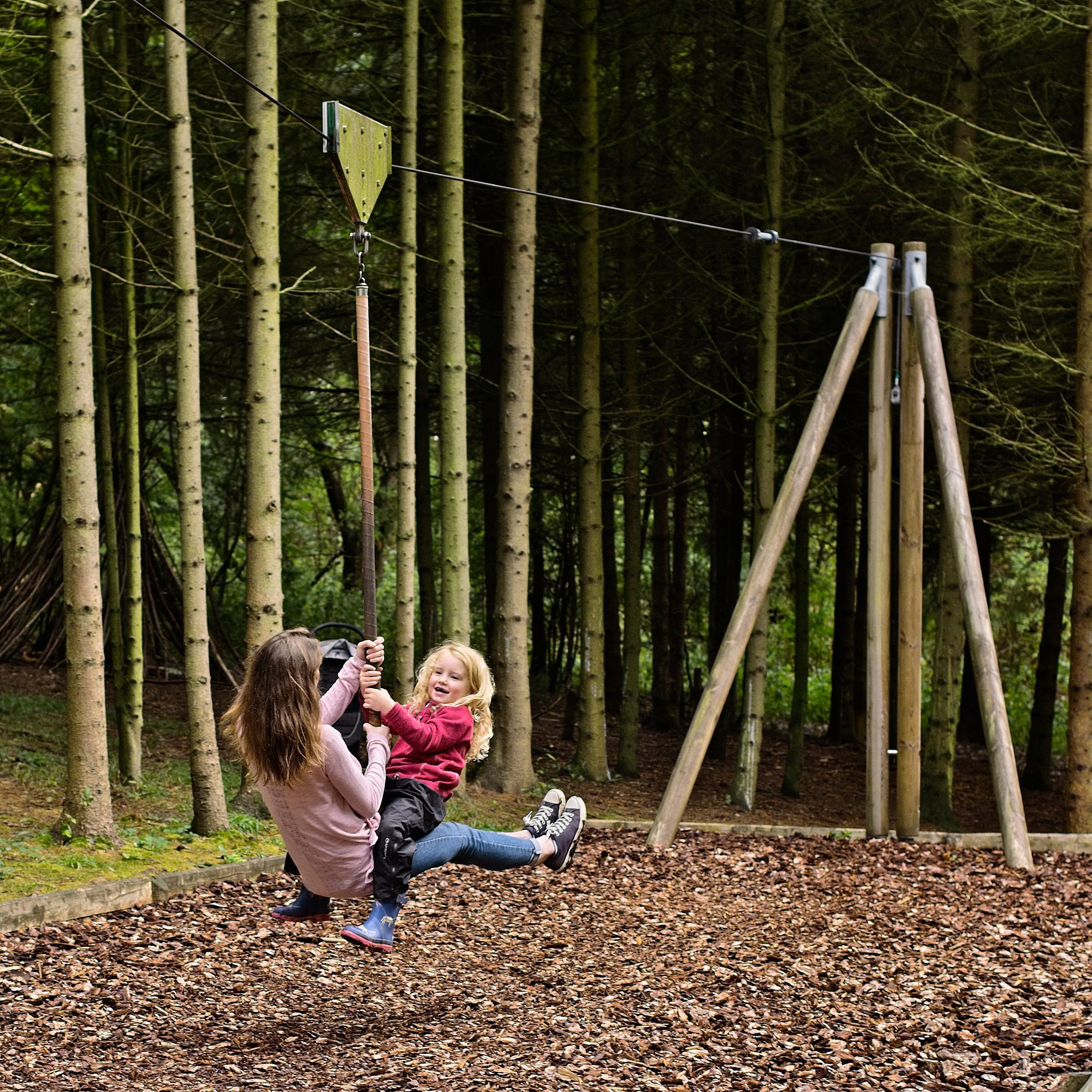 A woman is playfully playing on a zipline with a young girl in a wooded area with wooden structures in the background.