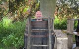 A smiling bald man sitting in what appears to be a large wooden barrel or tub outdoors. The barrel is situated among trees, suggesting it may be some kind of outdoor bath or spa experience in a natural setting.