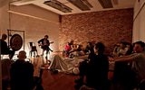 An indoor performance space with exposed brick walls and modern lighting fixtures. A musician is playing guitar on a small stage while audience members sit on the floor, creating an intimate concert atmosphere.