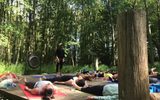 An outdoor yoga or meditation class in a forested setting. Several people are lying on yoga mats on a wooden platform. An instructor is standing, likely guiding the session. A large gong is visible, suggesting it may be used as part of the practice.