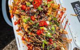 A plate filled with a colourful and flavourful dish, including fries, vegetables, and a meat topping. The food appears to be a savoury and spicy dish, likely a fusion of cuisines.