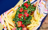 This image depicts a colourful food item, likely a specialty dish or snack, featuring chips or fried potatoes mixed with various chopped vegetables, possibly including kale or spinach, and topped with what appears to be a sauce or dressing.