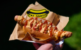 This image shows a hand holding a hot dog or sausage item that has been wrapped in a paper sleeve or container, with the 