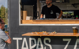 A food track with the word 'TAPAS' visible. There is a man standing behind the counter talking to another man in front of the van. There a plates of food on the shelf of the van.