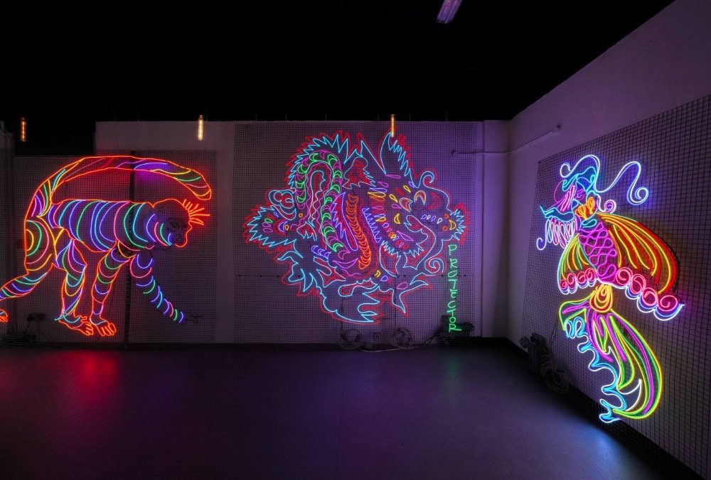 This image shows a dimly lit room with several large, neon-lit artworks on the walls. The artworks feature bold, dynamic designs in vibrant colors, creating a visually striking and immersive environment.