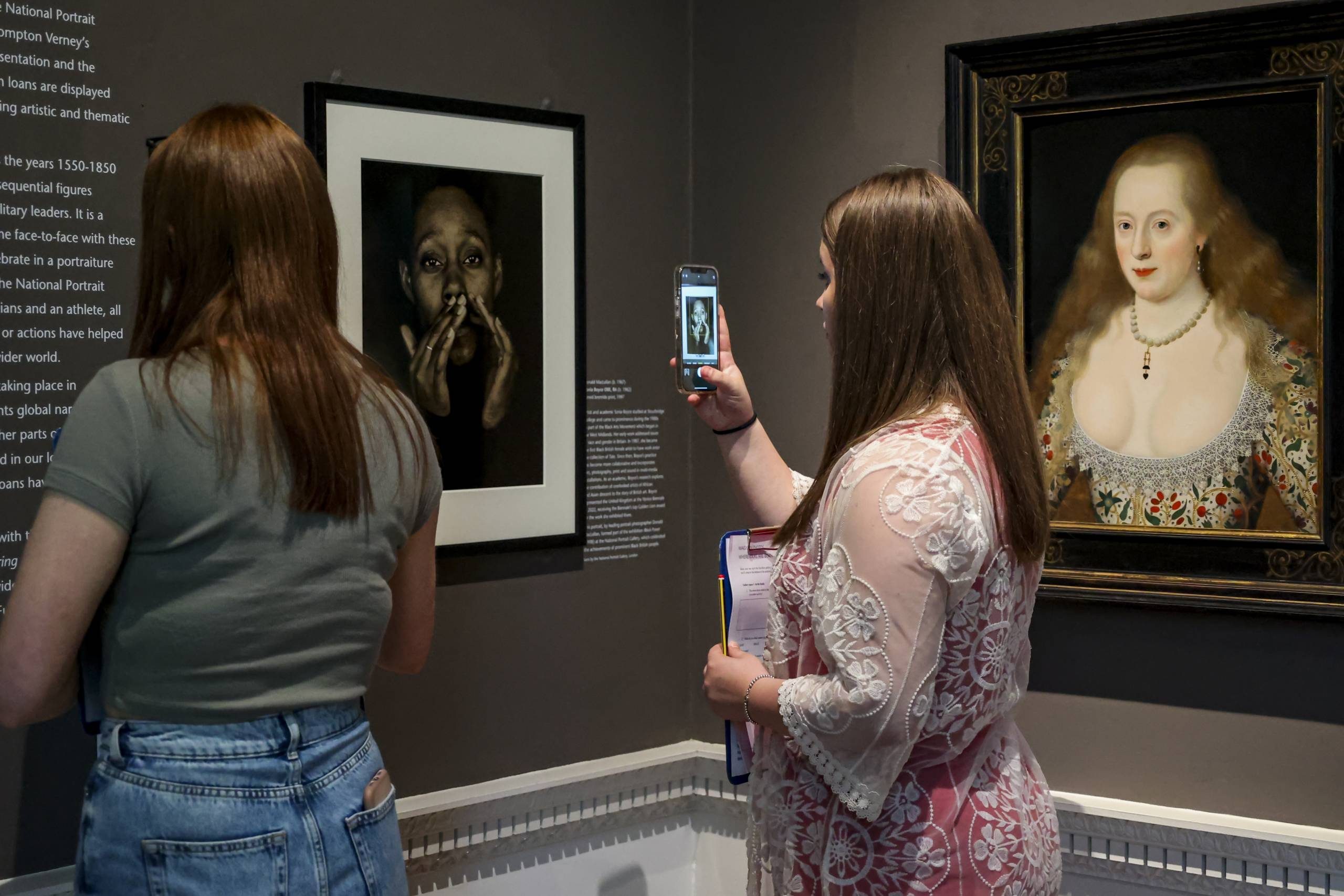 Two women in an art gallery looking at portraits. One woman is taking a photo with her phone of a black and white portrait of a person covering their mouth. Next to it is a classical portrait of a pale-skinned woman in ornate dress.