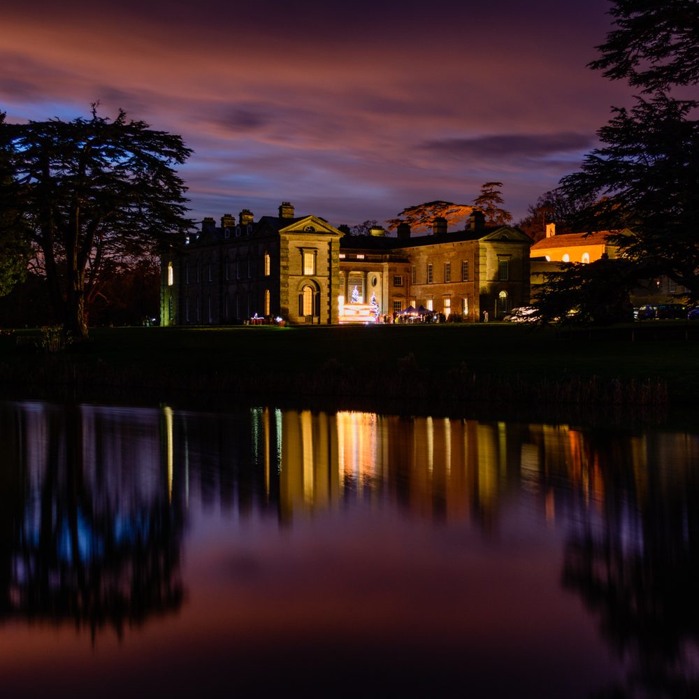 A night-time scene of a grand illuminated building, likely a manor or stately home, reflected in a still body of water. The sky is a mix of purple and orange hues, suggesting twilight or dawn. Trees frame the scene on both sides, and the building's lights create a warm glow that contrasts with the cooler tones of the sky and water.