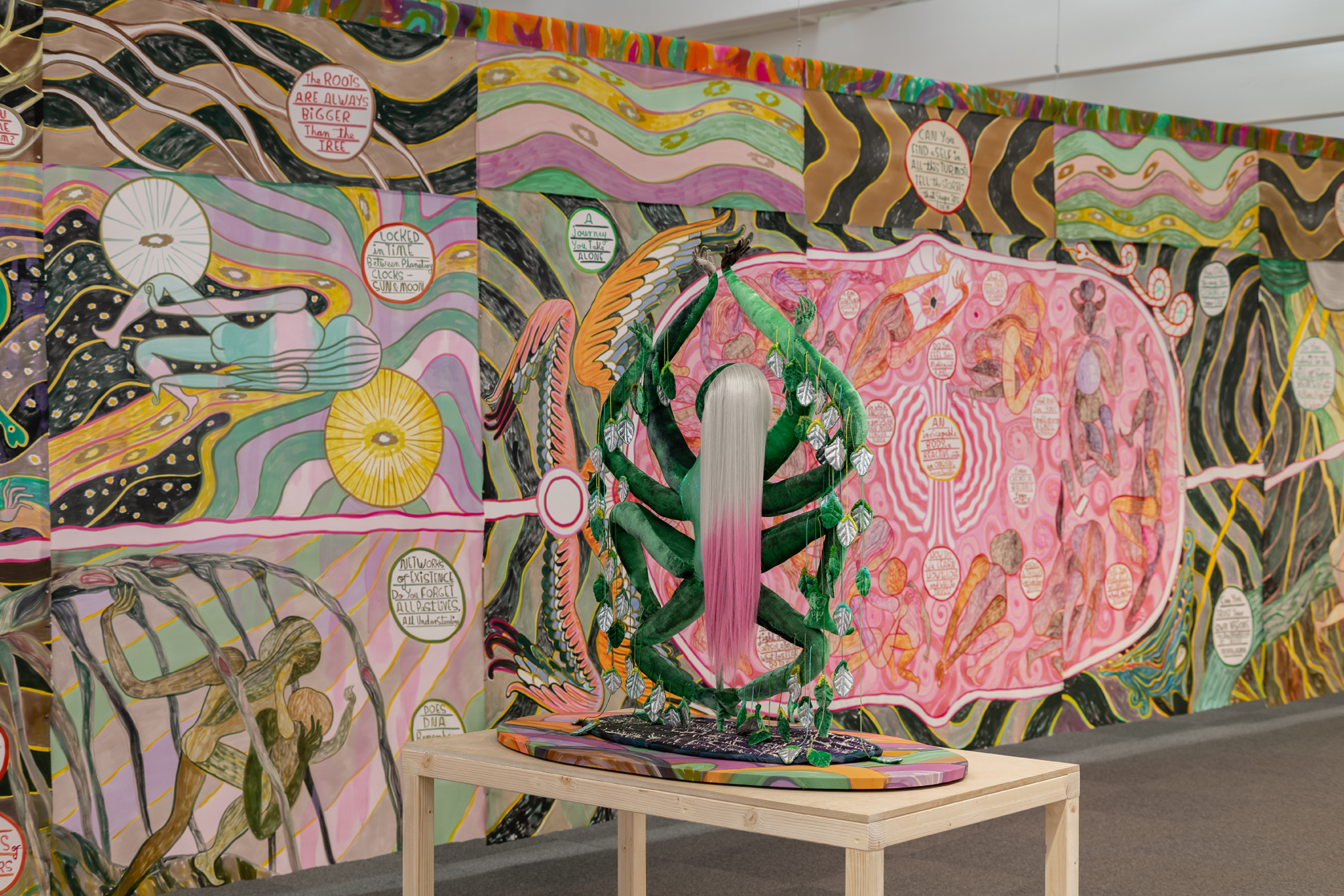 A large, vibrant mural covering the walls of an indoor space. The mural features a highly detailed, fantastical landscape with numerous surreal, symbolic elements. Swirling patterns, celestial imagery, and imaginary creatures create a sense of dreamlike, otherworldly atmosphere. A central, larger-than-life female figure with long, flowing pink hair appears to be the focal point of the composition.