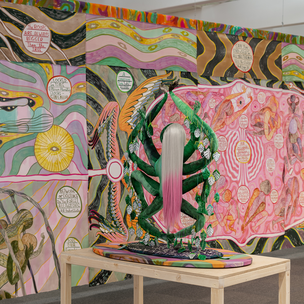 A large, vibrant mural covering the walls of an indoor space. The mural features a highly detailed, fantastical landscape with numerous surreal, symbolic elements. Swirling patterns, celestial imagery, and imaginary creatures create a sense of dreamlike, otherworldly atmosphere. A central, larger-than-life female figure with long, flowing pink hair appears to be the focal point of the composition.