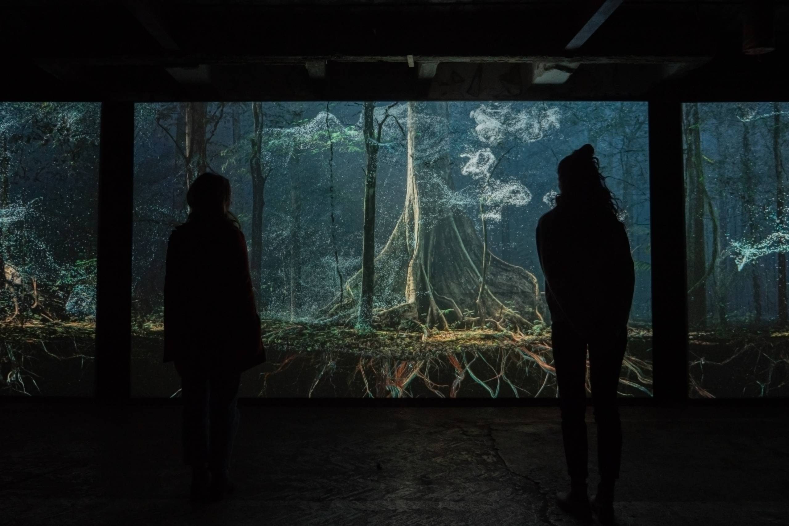 An immersive projection or display of a mystical forest scene, with two silhouetted figures viewing it. The forest has an ethereal blue-green glow with intricate root systems visible.