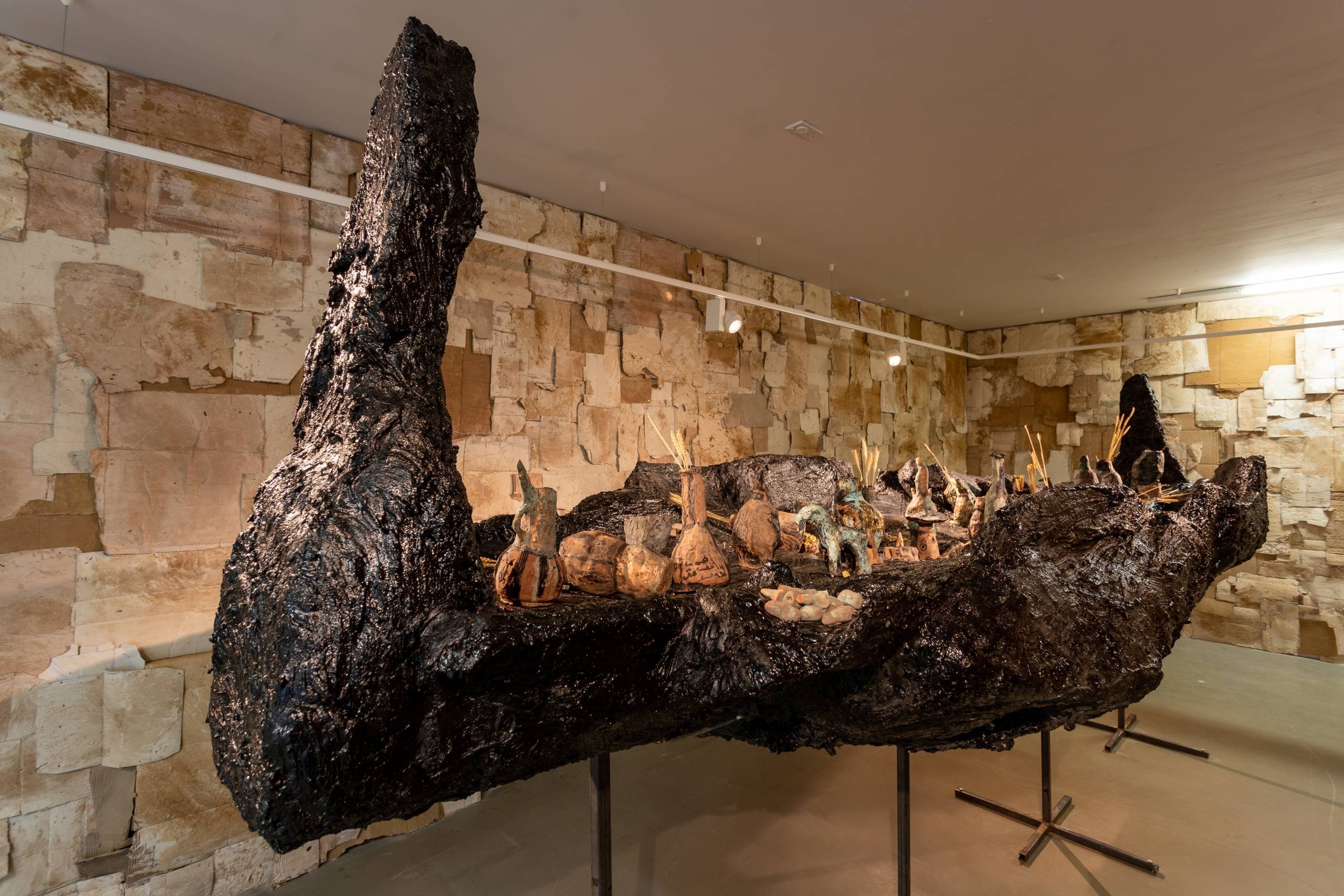 This image depicts a large, irregular-shaped sculpture made of dark, rough-textured material, possibly charred wood or a similar organic substance. The sculpture has an abstract, irregular form with many protruding elements. It is displayed on a raised platform in a spacious, gallery-like room with stone walls.