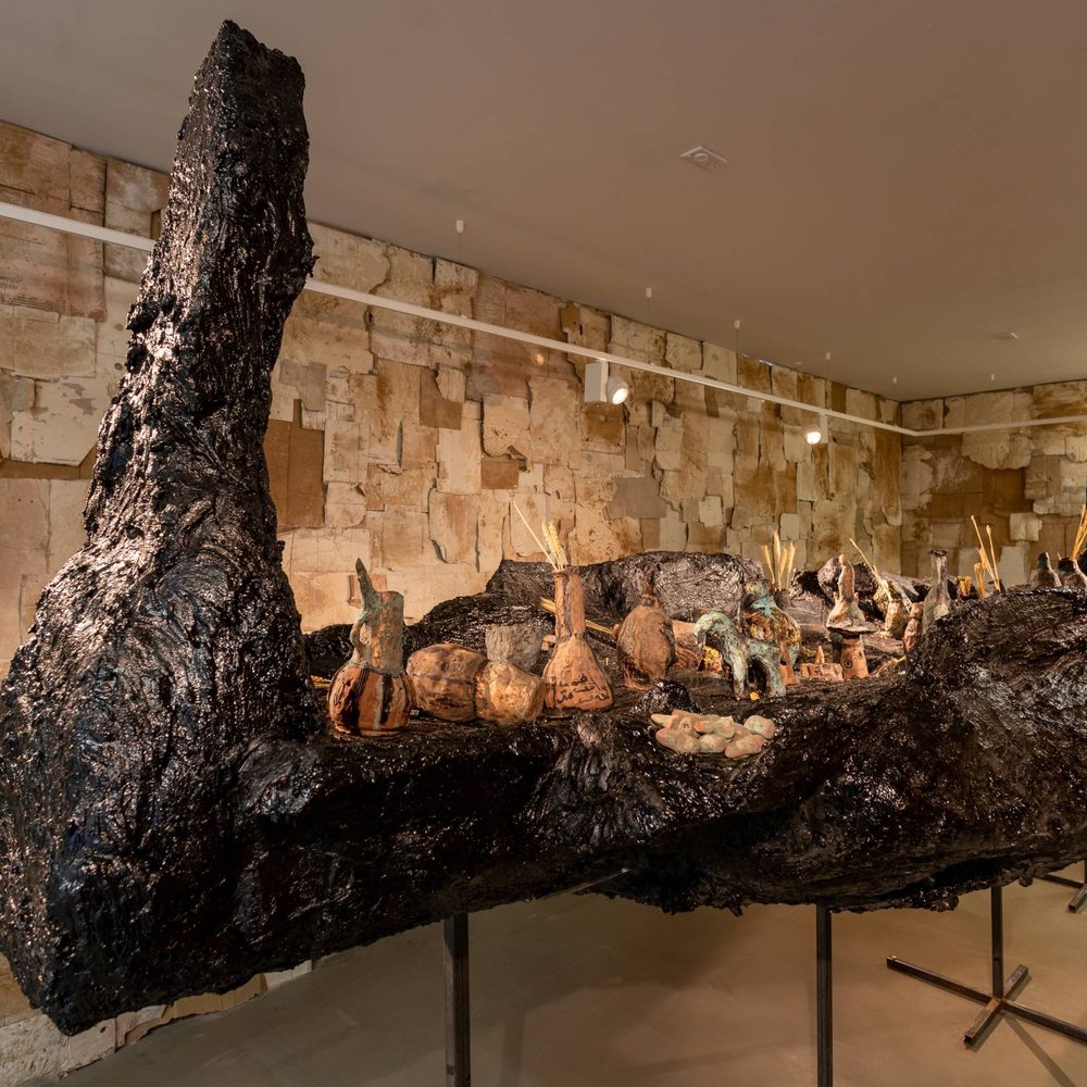 This image depicts a large, irregular-shaped sculpture made of dark, rough-textured material, possibly charred wood or a similar organic substance. The sculpture has an abstract, irregular form with many protruding elements. It is displayed on a raised platform in a spacious, gallery-like room with stone walls.