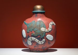 Install View, Commodities by Renee So. Photo © Compton Verney and Jamie Woodley (1) A recreation of a Chinese snuff bottle with coral-pink base colour decorated with turquoise and white painted designs resembling stylized elephants or animals, topped with a brass cap and turquoise collar, displayed against a reddish-brown background.