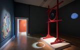 Contemporary museum gallery with dark teal walls displaying a large red sculptural installation resembling cuju goal posts. Holding a magic mirror, projecting an image of cuju onto the wall. There is a mosaic image of two women playing cuju on the opposite wall and you can see this room is, connected to an orange-walled gallery space beyond.