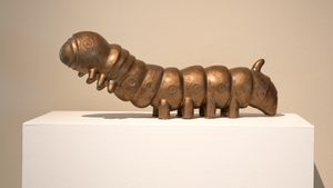 Install View, Commodities by Renee So. Photo © Compton Verney and Jamie Woodley (20) A bronze or copper-colored sculptural silkworm displayed on a white pedestal against a neutral beige background. The segmented insect sculpture features a rounded head with circular markings and multiple body segments, each with small circular details. The silkworm has small legs protruding from its underside and has a metallic, patinated finish.