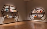 A museum gallery corner featuring two distinctive wall-mounted display cases with wooden frames. On the left is a hexagonal shelving unit containing various ceramic vessels and pottery in different colours including turquoise, pink, cream, and black. On the right is a circular display case housing what appears to be perfume bottles and cosmetic containers, including items labelled 