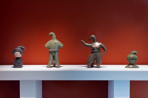 Install View, Commodities by Renee So. Photo © Compton Verney and Jamie Woodley (22) Four bronze or metal sculptural figures arranged on a white display shelf against a rich red-orange wall. The sculptures appear to be stylized human forms with distinctive curved, heads. They vary in size and patina, ranging from greenish to dark bronze finishes. The figures have simplified, abstract body forms.