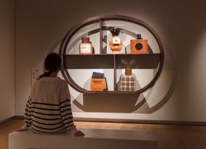 Install View, Commodities by Renee So. Photo © Compton Verney and Jamie Woodley (3) Museum visitor in striped sweater sits on bench viewing a circular wooden display case containing vintage perfume bottles and containers, including items labeled