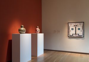 Install View, Commodities by Renee So. Photo © Compton Verney and Jamie Woodley (7) Museum gallery interior showing two decorative vessels on white pedestals against an orange accent wall, with a framed geometric artwork featuring a face-like design hanging on the adjacent neutral wall.