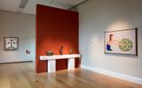 Museum gallery space with white walls and wooden floors, featuring ancient pottery vessels on a white display table against an orange accent wall, with geometric artworks including a face-like design and a figure with circular motif hanging on the walls.