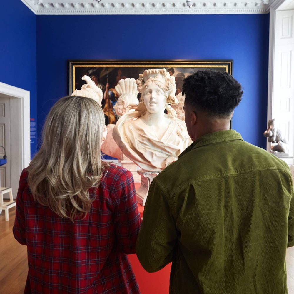 Rear view of two people viewing a marble bust sculpture in a museum with royal blue walls and wooden floors.