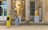 Two visitors view a large outdoor golden sculpture shaped like an ornate trophy with eyes and a face, displayed outside a historic stone building.