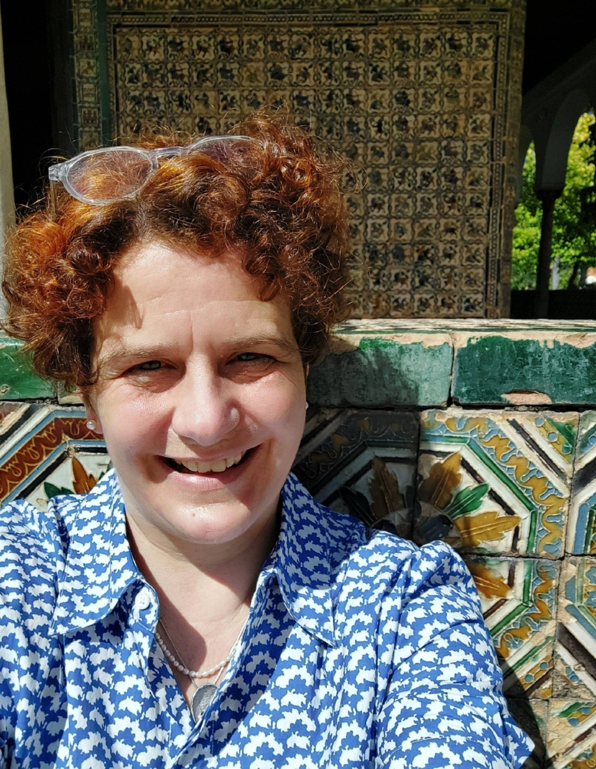 Portrait of a woman with short curly red hair, wearing a blue patterned shirt, outdoors in bright sunlight.