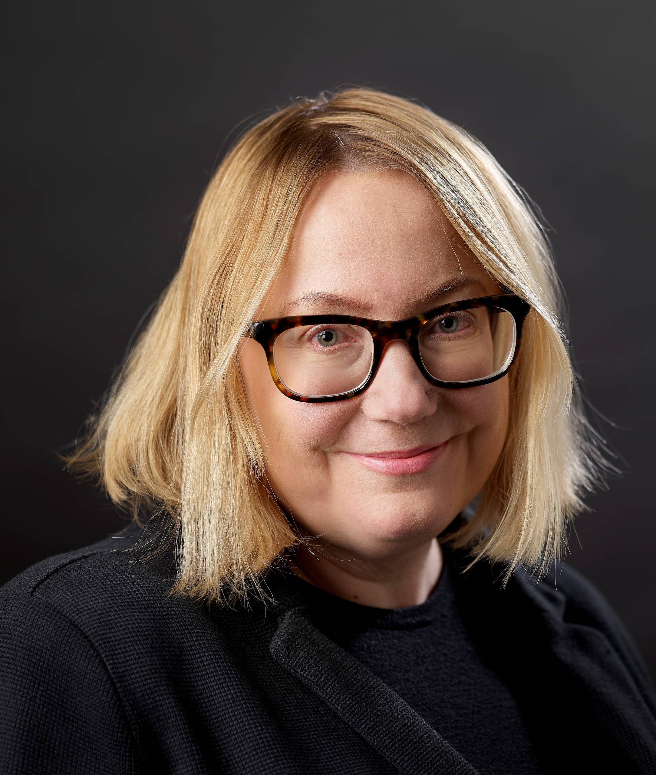 Portrait of a woman with blonde hair and glasses, wearing black, against a dark background.