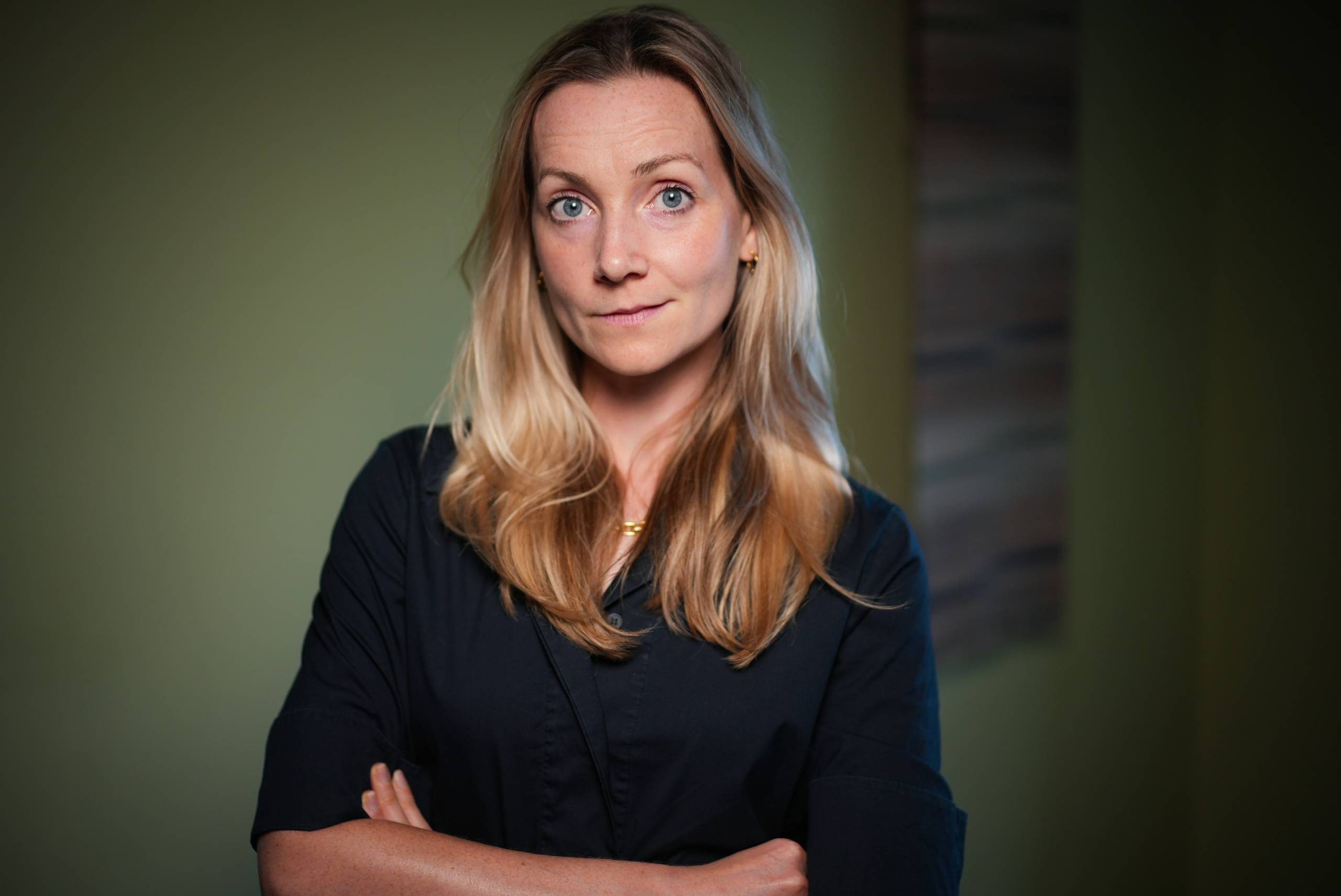 Portrait of a woman with long blonde hair, wearing a dark top, against a blurred green background.