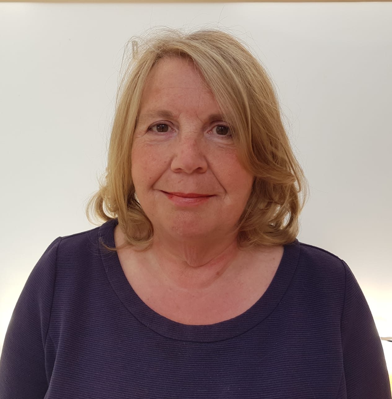 Portrait of a woman with shoulder-length blonde hair, wearing a dark purple top, against a plain light background.